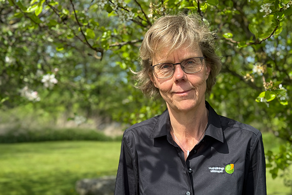 Kristina Lidfeldt står utomhus framför ett träd med blommande grenar. Hon har kort, ljusbrunt hår, glasögon och bär en svart skjorta med Hushållningssällskapets logotyp. Hon tittar lugnt mot kameran, och i bakgrunden syns grönskande natur i solsken.