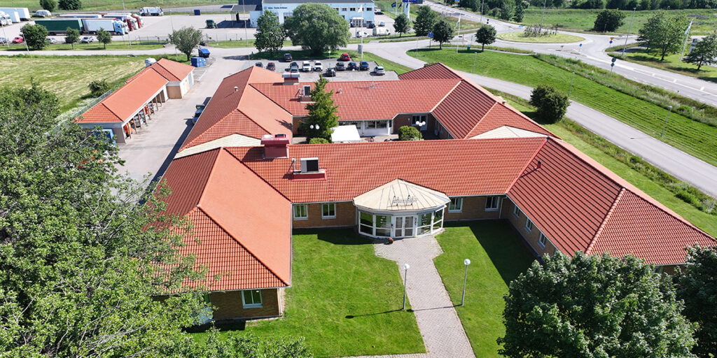Drönarfoto över Agrocenter utanför Falkenberg, en enplansbyggnad med rött tegeltak och ljus tegelfasad. Byggnaden har en H-formad struktur med en glasentré i mitten, omgiven av välskötta gräsmattor och gångvägar. I bakgrunden syns en parkeringsplats, vägkorsningar och omgivande grönområden.