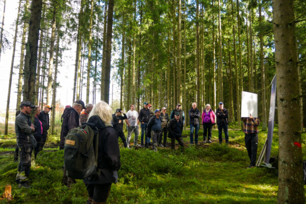 Människor i grupp som står i skogen. De lyssnar på en man med skylt.